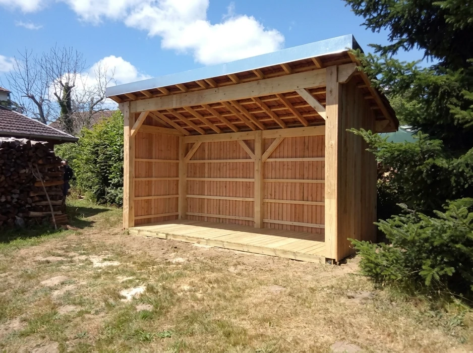 Maison individuelle neuve en construction avec façade crème, volets blancs et toiture tuiles rouges, située sur terrain en pente boisée